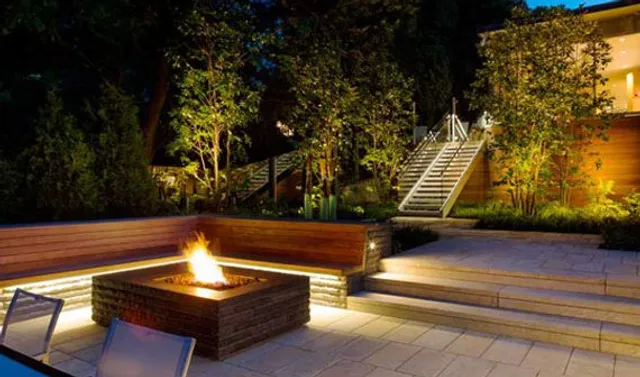 Terraced patio with a square fire pit, bench seating, and step lighting at night