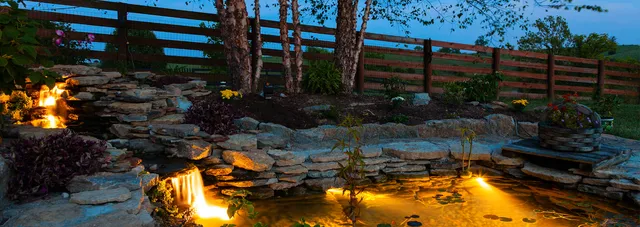 Backyard pond and waterfall lit at night beside a paver terrace